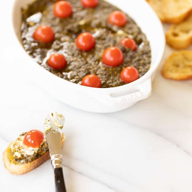 White oval baking dish on marble surface, filled with easy appetizer dip and cherry tomatoes on top, spoon and crostini to the side.