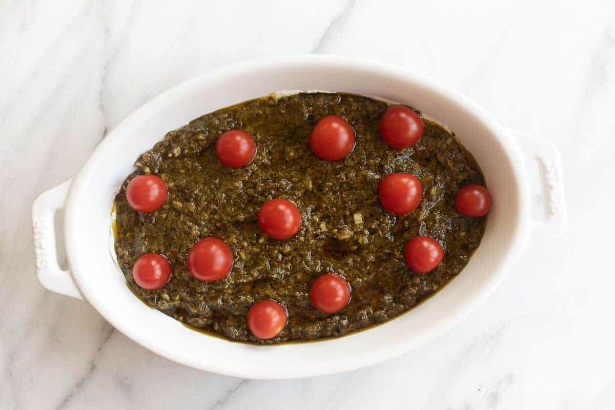 White oval baking dish on marble surface, filled with pesto dip and cherry tomatoes on top.