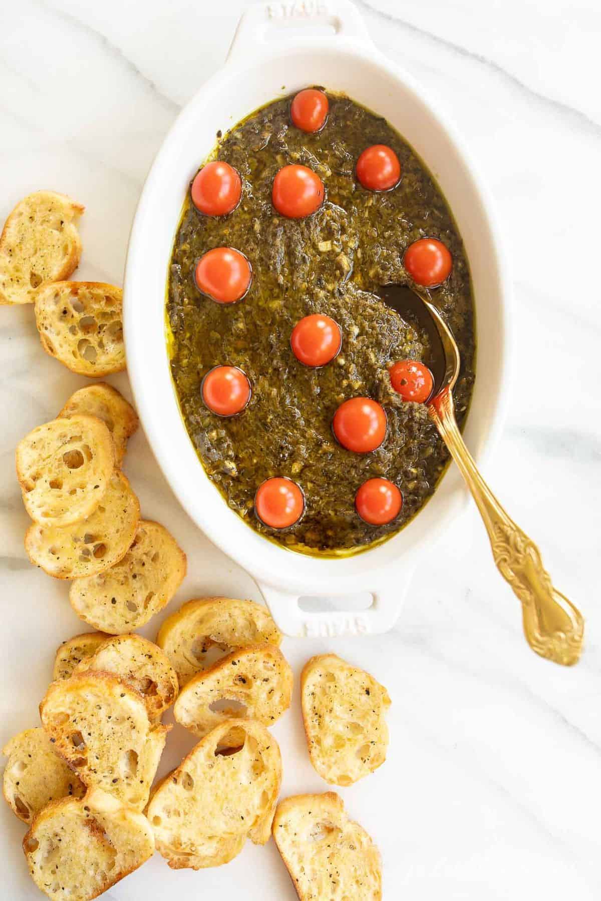 White oval baking dish on marble surface, filled with pesto dip and cherry tomatoes on top, spoon and crostini to the side.