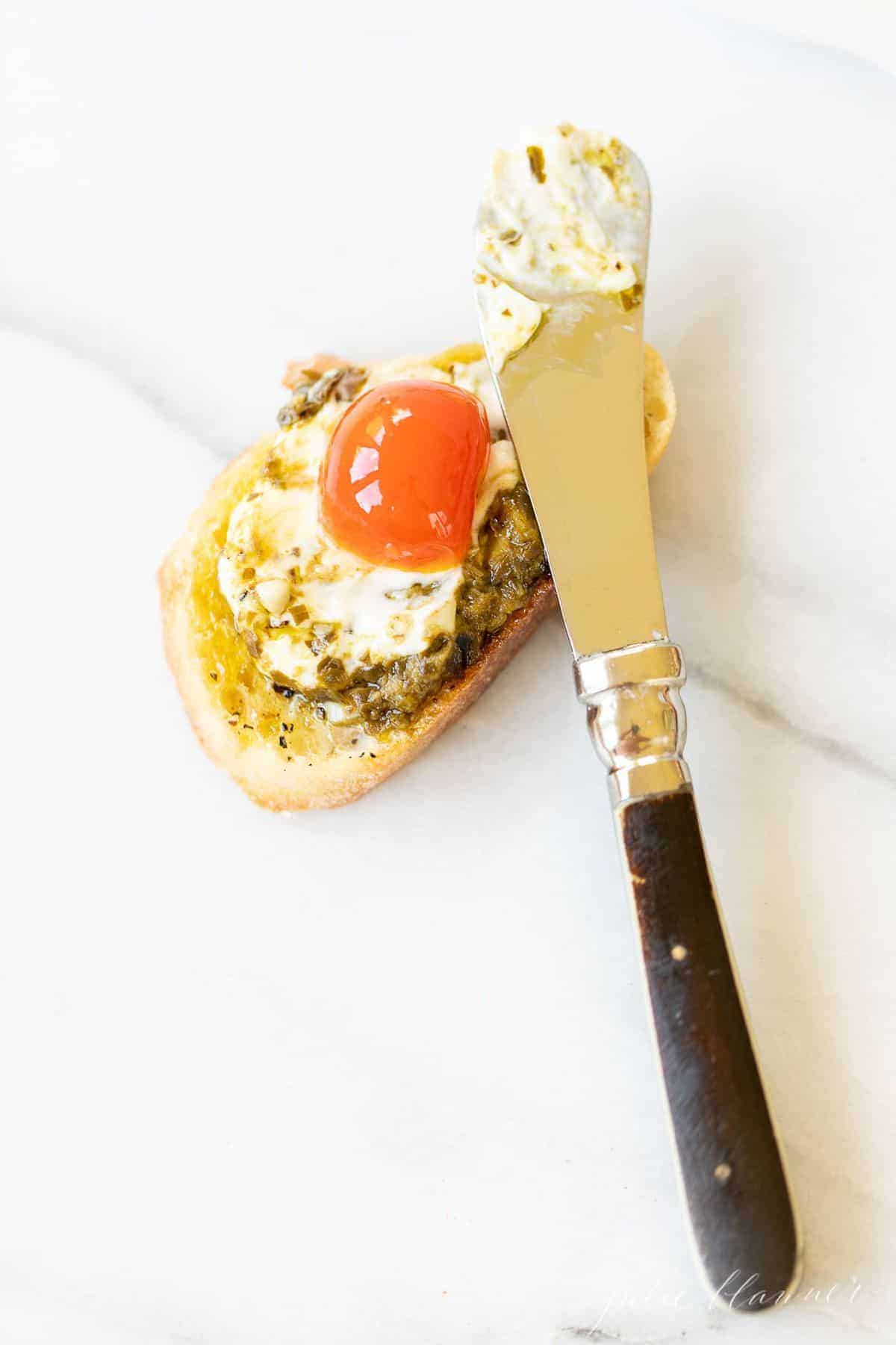 Marble surface, single slice of crostini topped with pesto cheese dip and a cherry tomato, spreader on top.