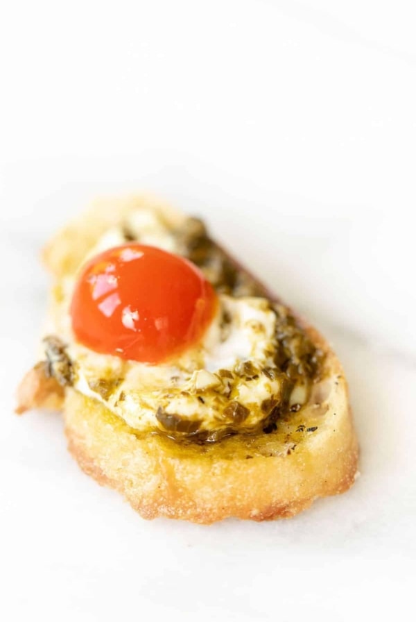 Marble surface, single slice of crostini topped with pesto cheese dip and a cherry tomato.