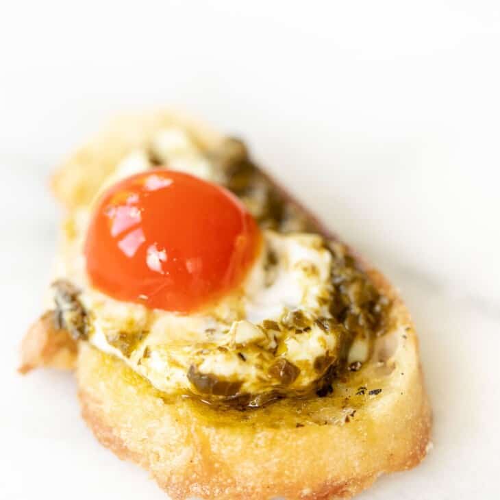Marble surface, single slice of crostini topped with pesto cheese dip and a cherry tomato.