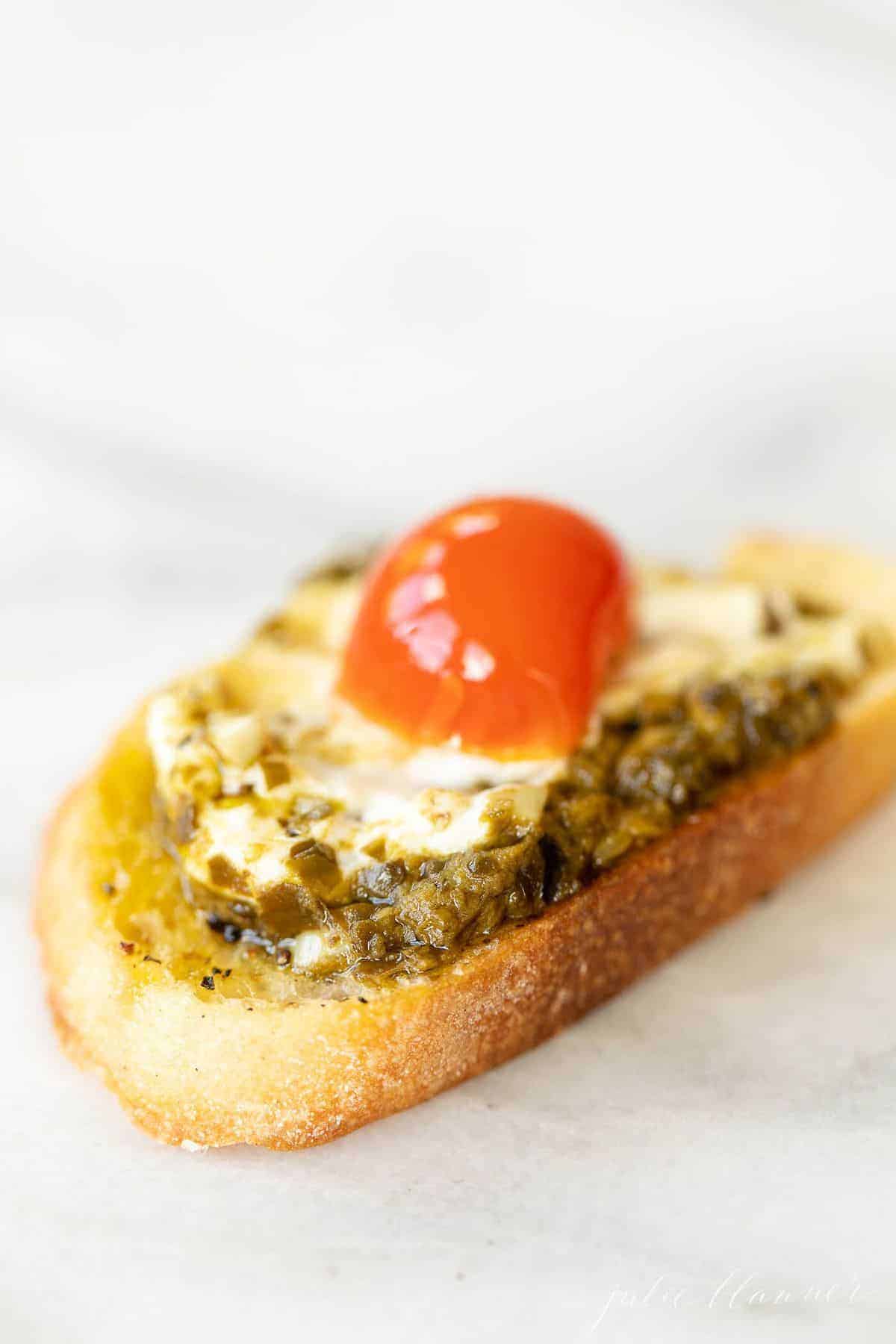 Marble surface, single slice of crostini topped with pesto cheese dip and a cherry tomato.
