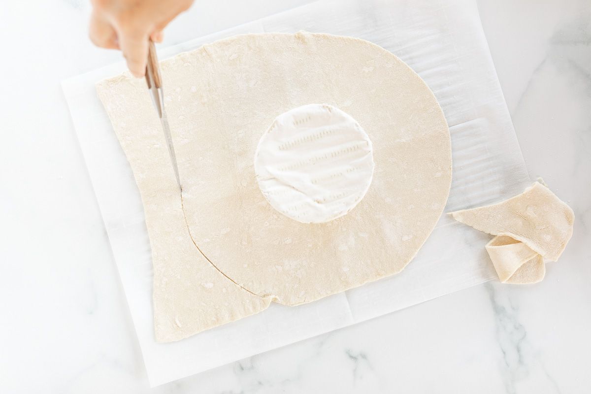 A sheet of puff pastry being cut under a round of brie.