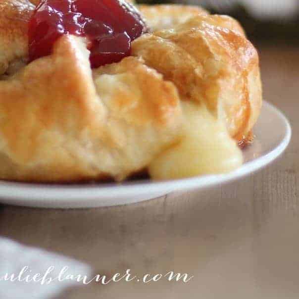 A close up of baked brie in puff pastry on a white plate, topped with jam and brie oozing out from the pastry.