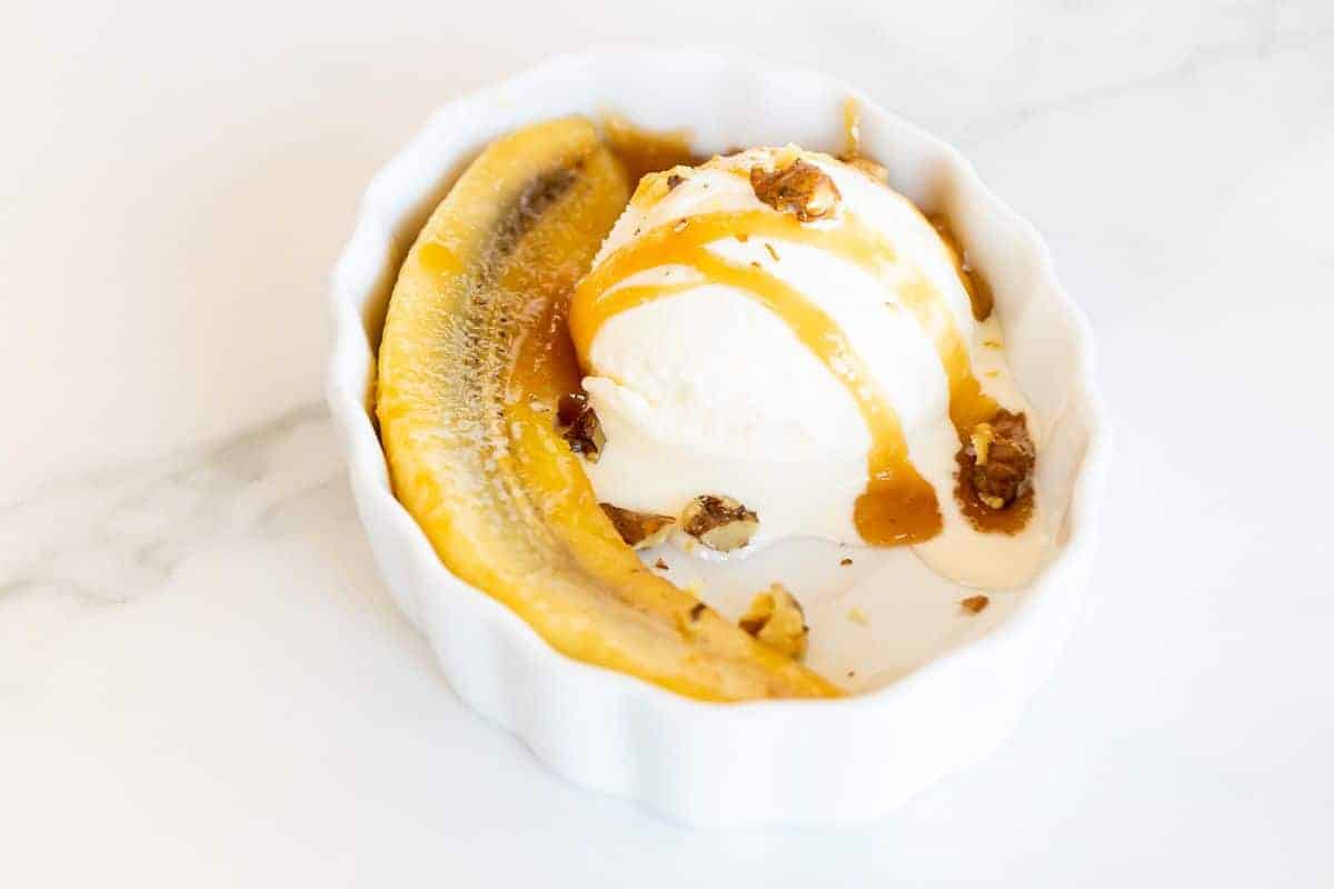 An oval ramekin filled with a bananas foster recipe with a scoop of ice cream drizzled in caramel.