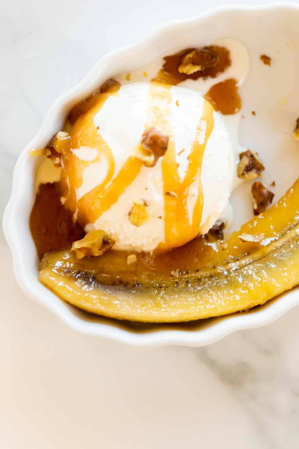 An oval ramekin filled with a cooked banana and a scooper of ice cream with caramel sauce.