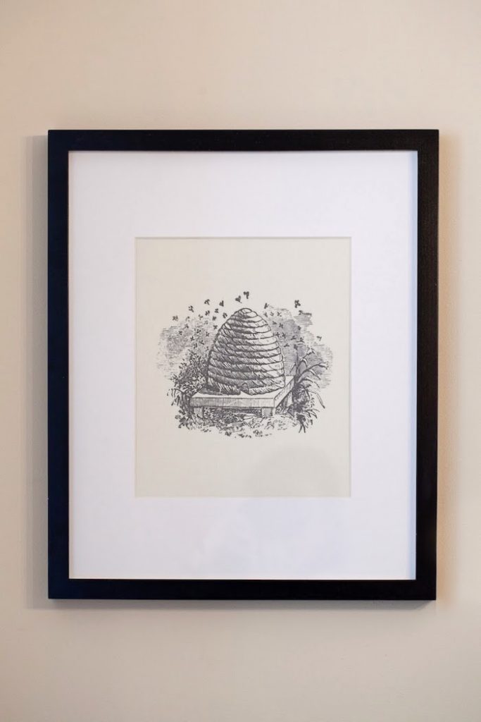 Framed art print of small beehive on wooden stand, with bees buzzing around it.