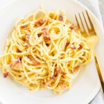 A white plate with a serving of spaghetti carbonara, gold fork to the side