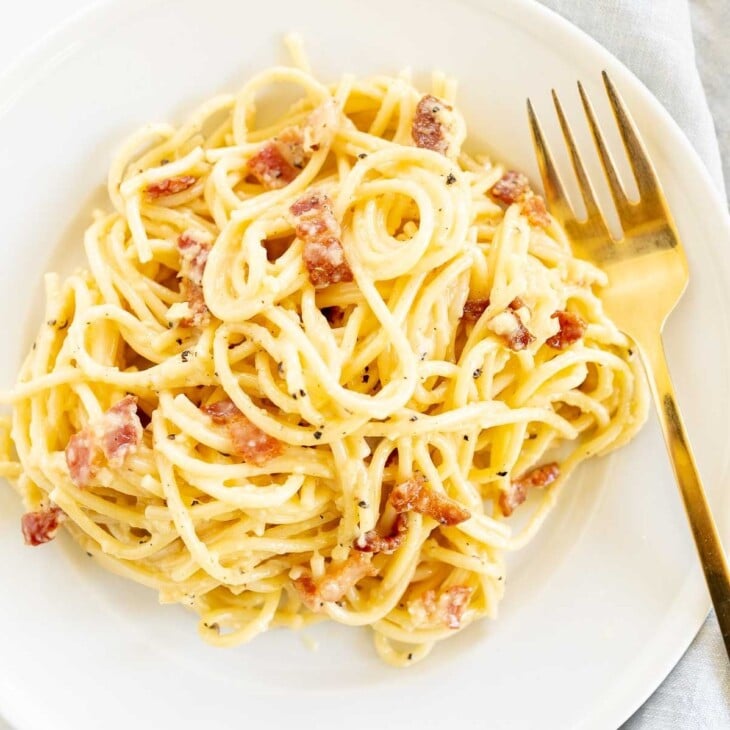 A white plate with a serving of spaghetti carbonara, gold fork to the side