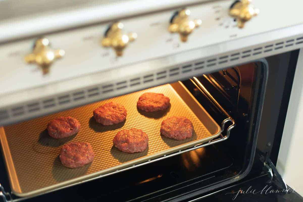 Six beef patties on a gold baking sheet in the oven. #sliders #easysliders.