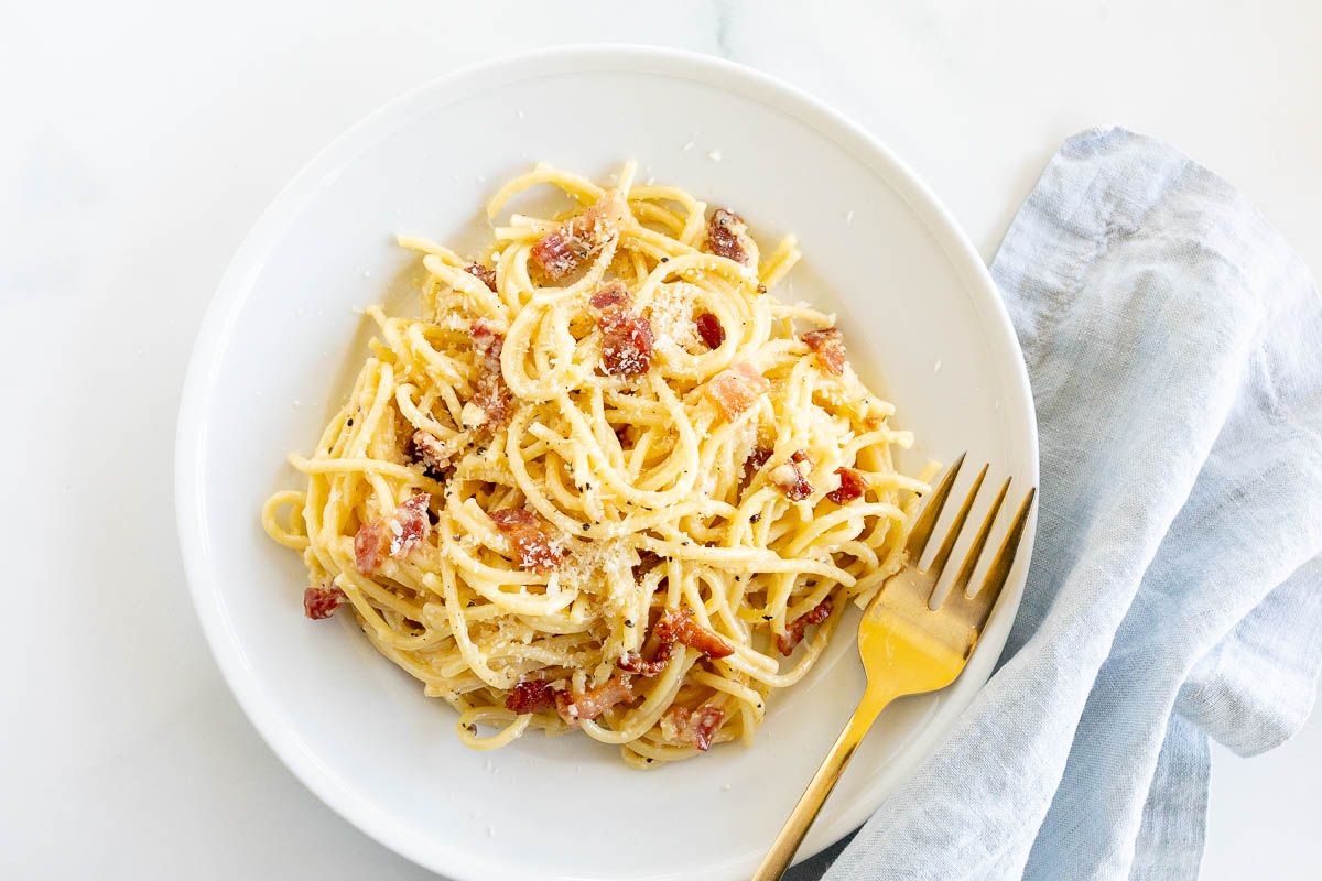 A white plate with a serving of spaghetti carbonara, gold fork to the side