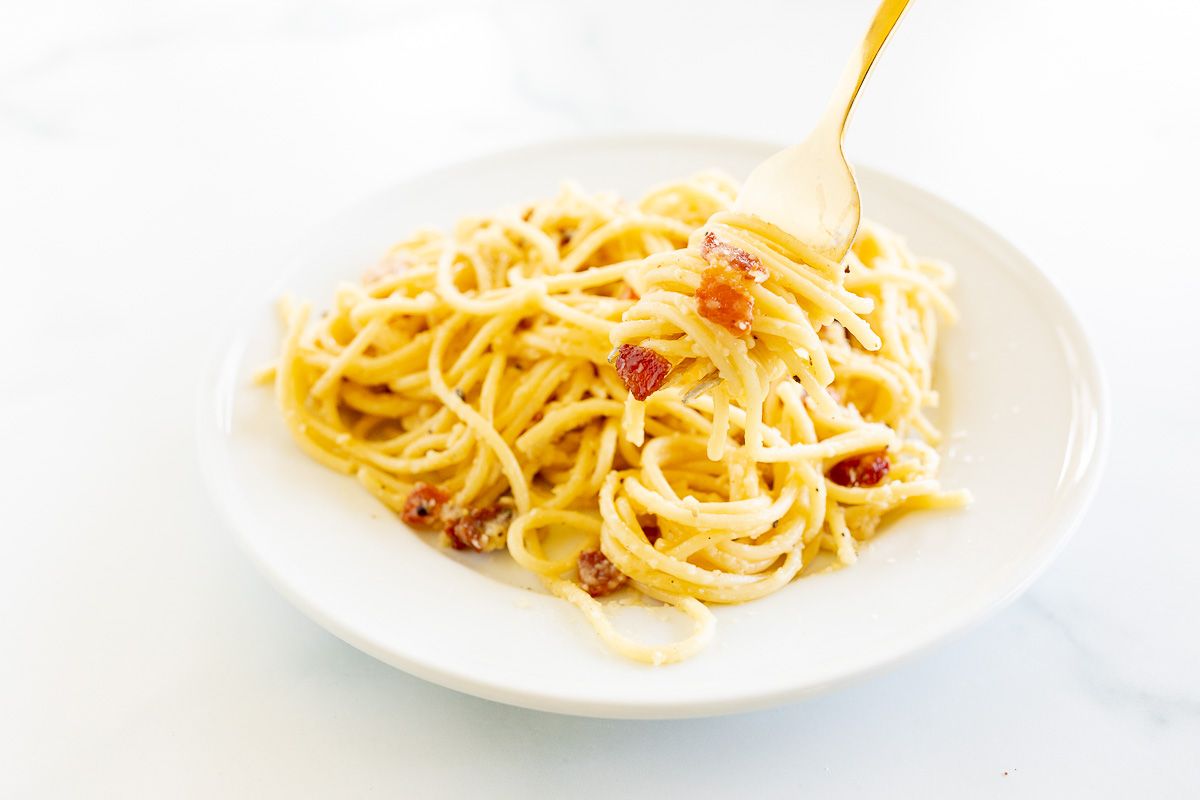 A white plate with a serving of spaghetti carbonara, gold fork sticking out