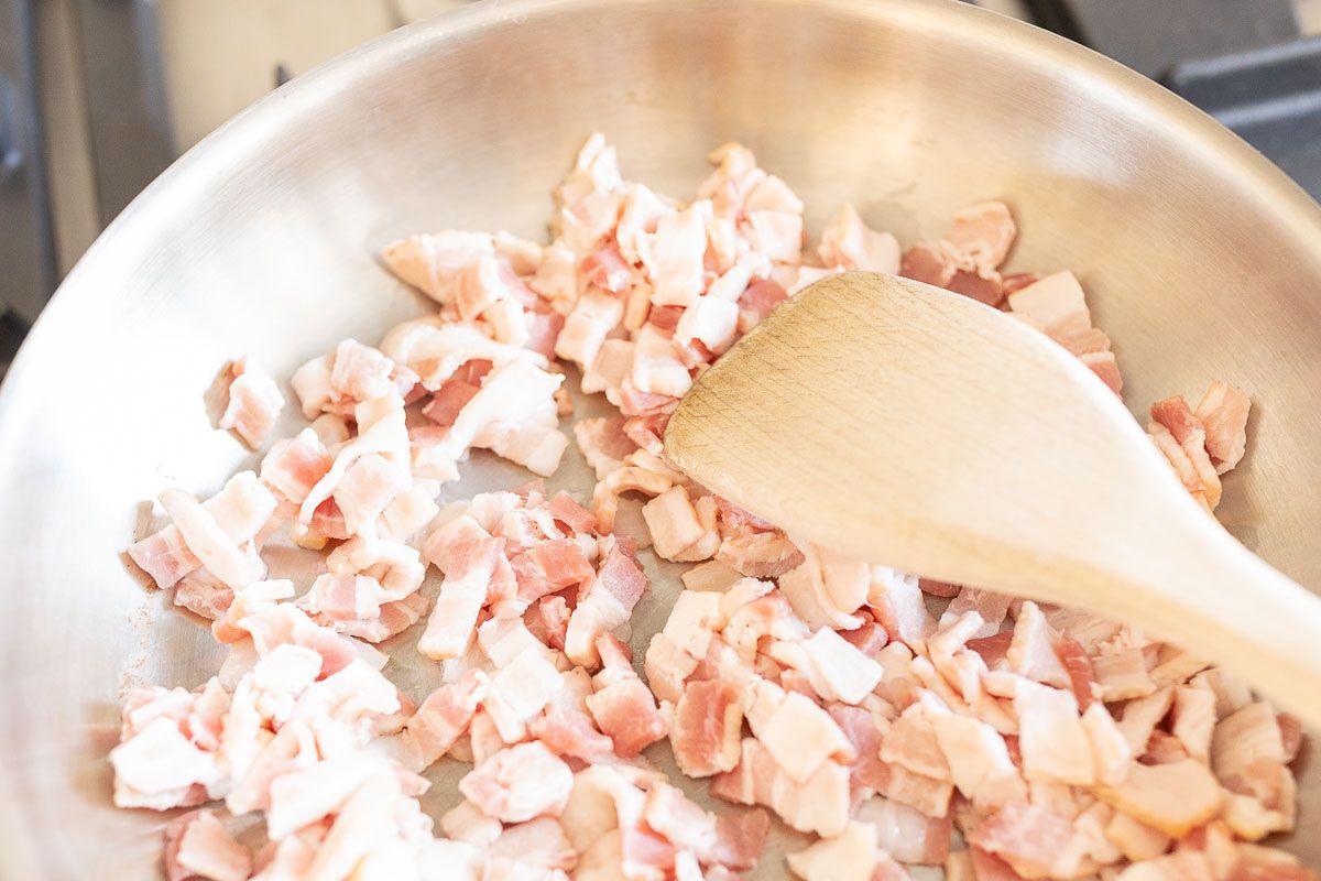 A silver pan with chopped bacon being cooked with a wooden spoon