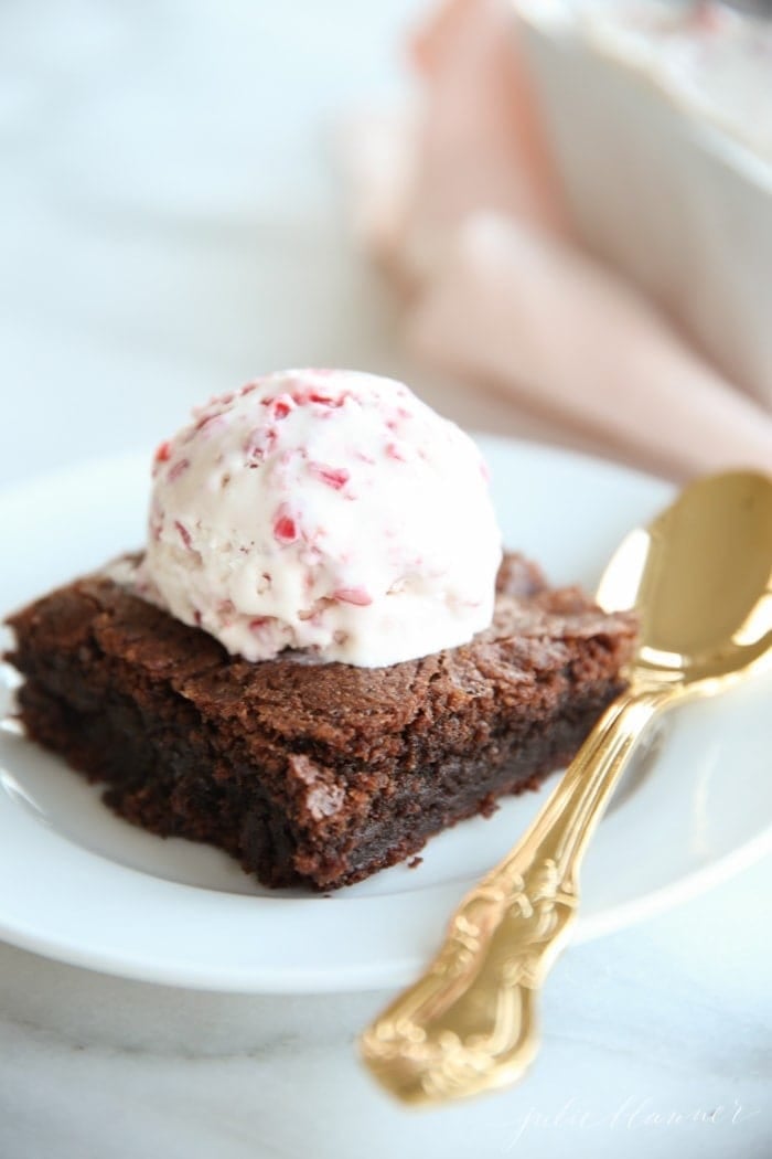 A homemade brownie on a plate, with a scoop of peppermint ice cream on top. Gold spoon rests to the side. 