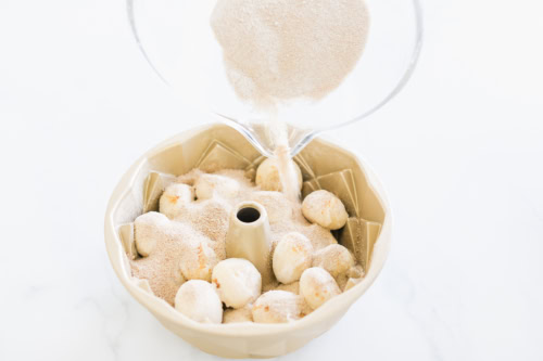 Monkey bread dough balls in a bundt pan are gracefully coated with sugar from a pouring container.