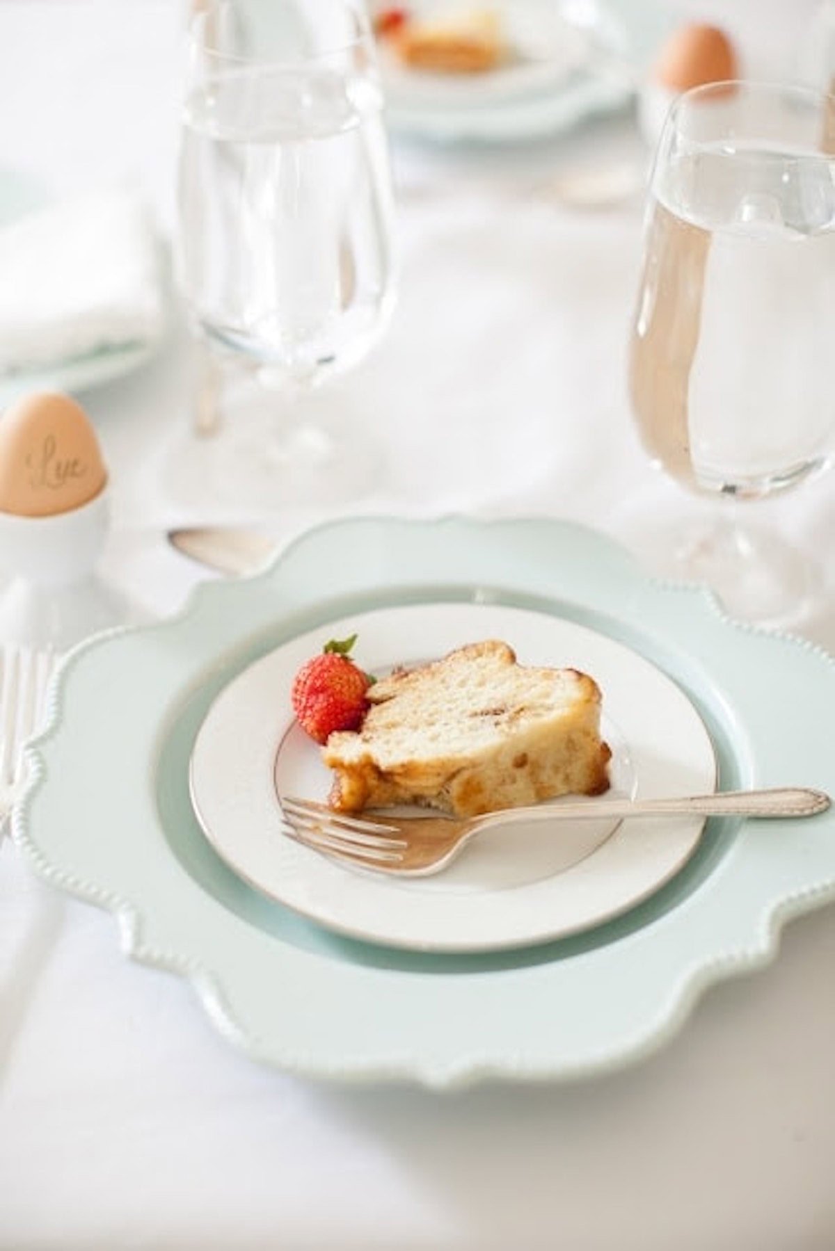 Slice of cake with a strawberry and a fork on a white plate, placed on a larger light blue plate; water glasses, soft-boiled egg in an egg cup, and overnight monkey bread are in the background on a white tablecloth.
