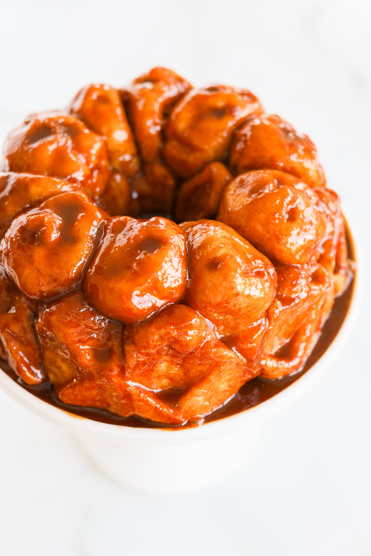 A close-up of homemade monkey bread in a white bowl showcases golden-brown dough pieces drenched in a shiny caramel glaze.