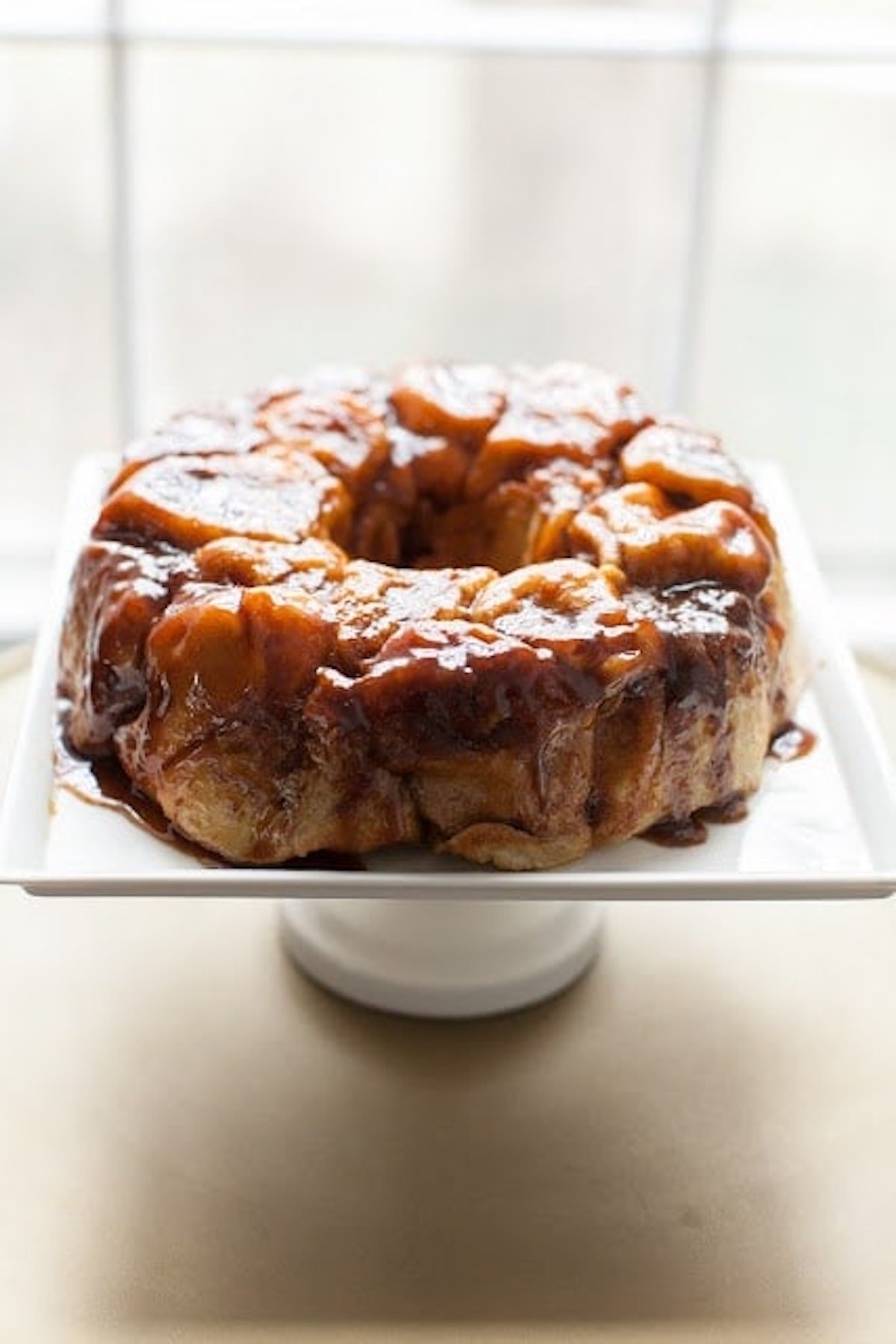 A caramel-glazed overnight monkey bread sits on a white square cake stand in front of a blurred window.