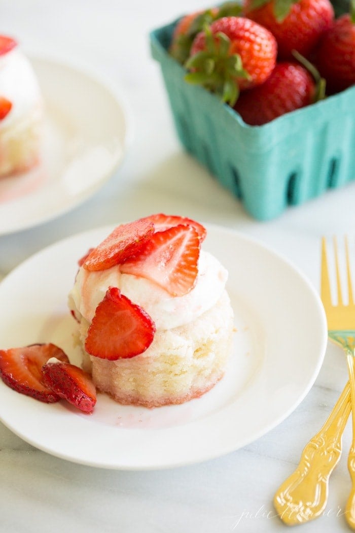 Strawberry shortcake on a white plate, turquoise box of fresh strawberries in background, gold flatware to the side.