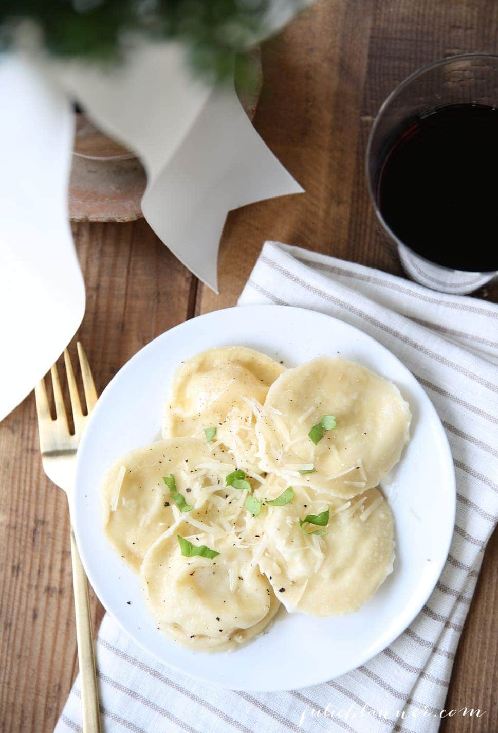 A plate of homemade ravioli on a striped napkin, wine and gold flatware surrounding.