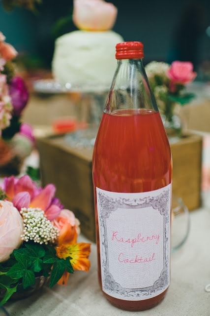 A close up of a raspberry cocktail in a clear glass bottle, with a fabric printed label.