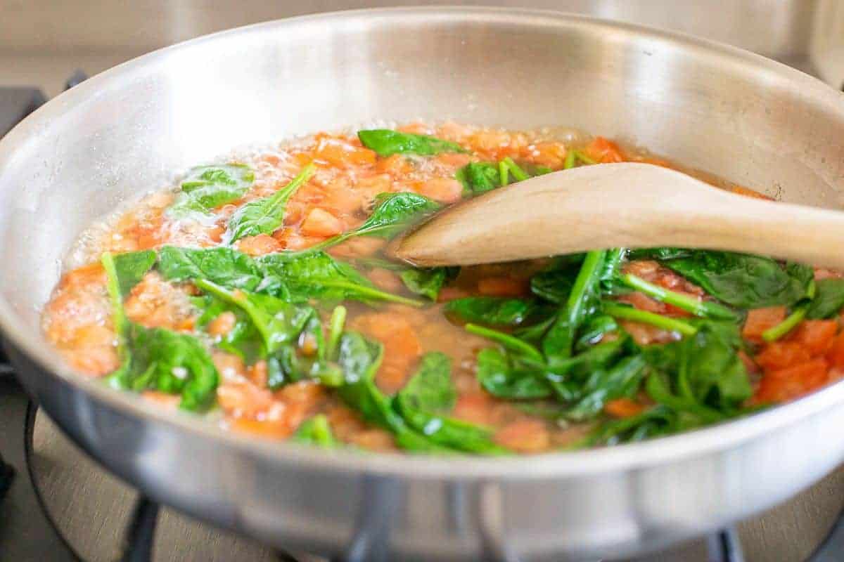 Spinach leaves cooking in a red sauce in a silver pan on a stove.