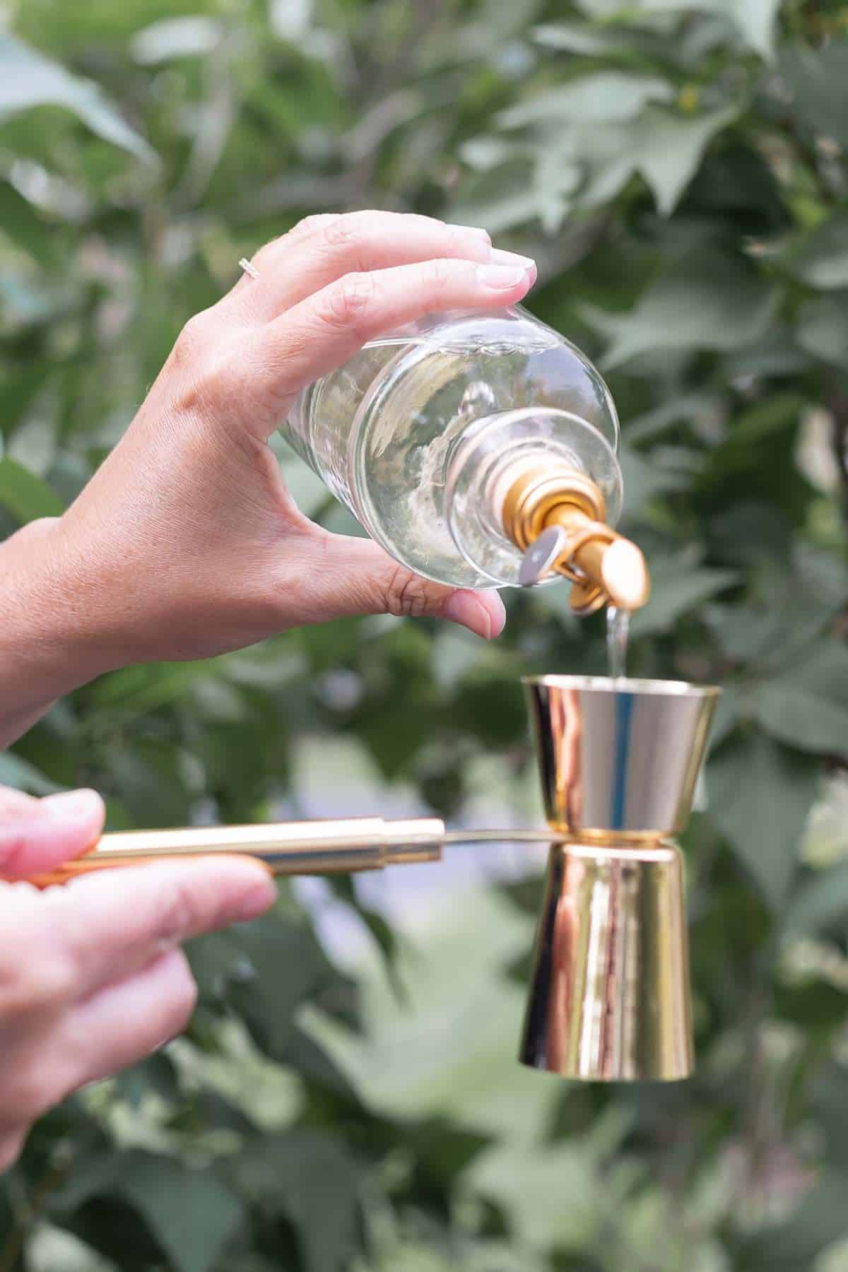 A hand pouring a glass bottle full of mixer into a copper shot measurer.