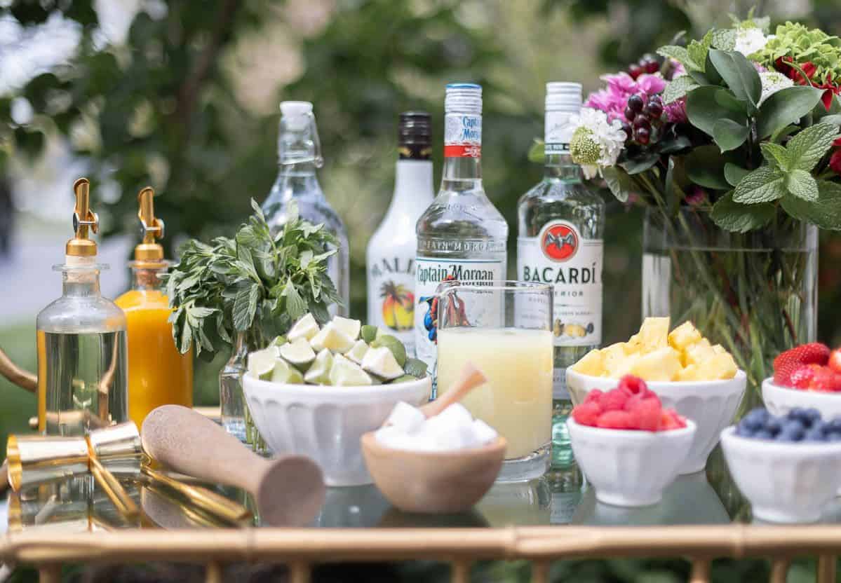 A glass and brass bar cart set up with a mojito bar outdoors, various fruits and limes in bowls beside alcohol bottles.