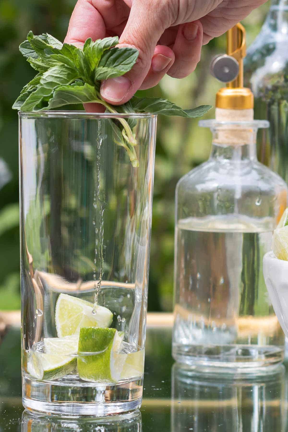 A mojito bar set up on a gold and glass bar cart, a hand reaching over to place fresh mint in a glass.