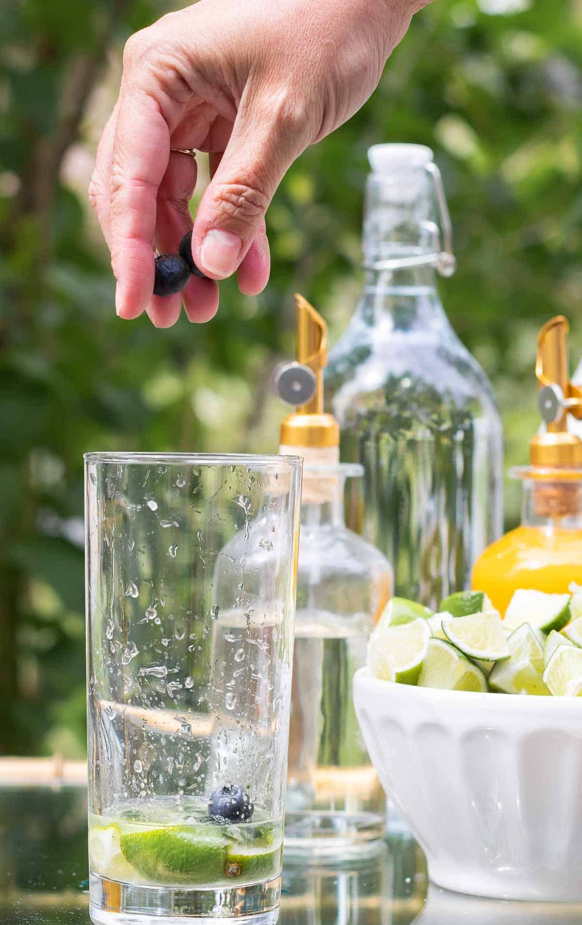 A mojito bar set up on a gold and glass bar cart, a hand reaching over to place blueberries in a glass.