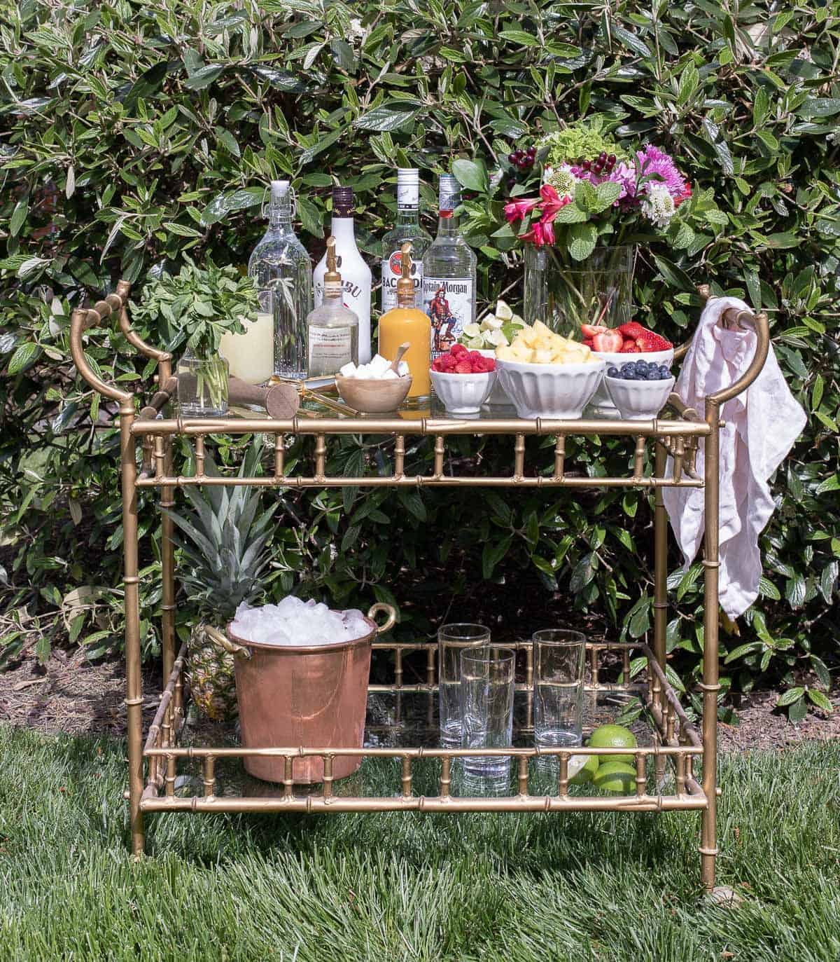 A glass and brass bar cart set up with a mojito bar outdoors.