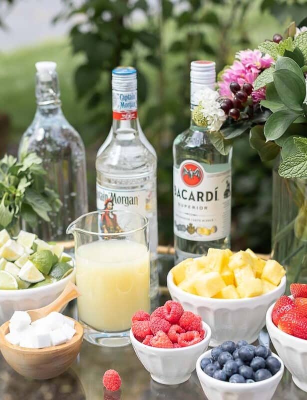 A glass and brass bar cart set up with a mojito bar outdoors, various fruits and limes in bowls beside alcohol bottles.