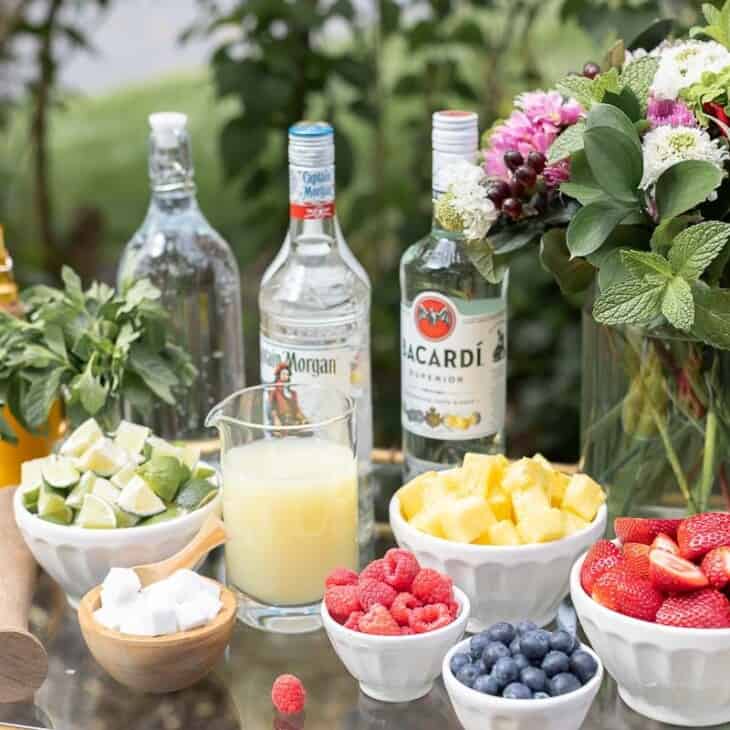 A glass and brass bar cart set up with a mojito bar outdoors, various fruits and limes in bowls beside alcohol bottles.