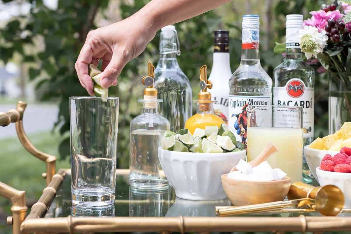 A mojito bar set up on a gold and glass bar cart, a hand reaching over to place a lime in a glass.