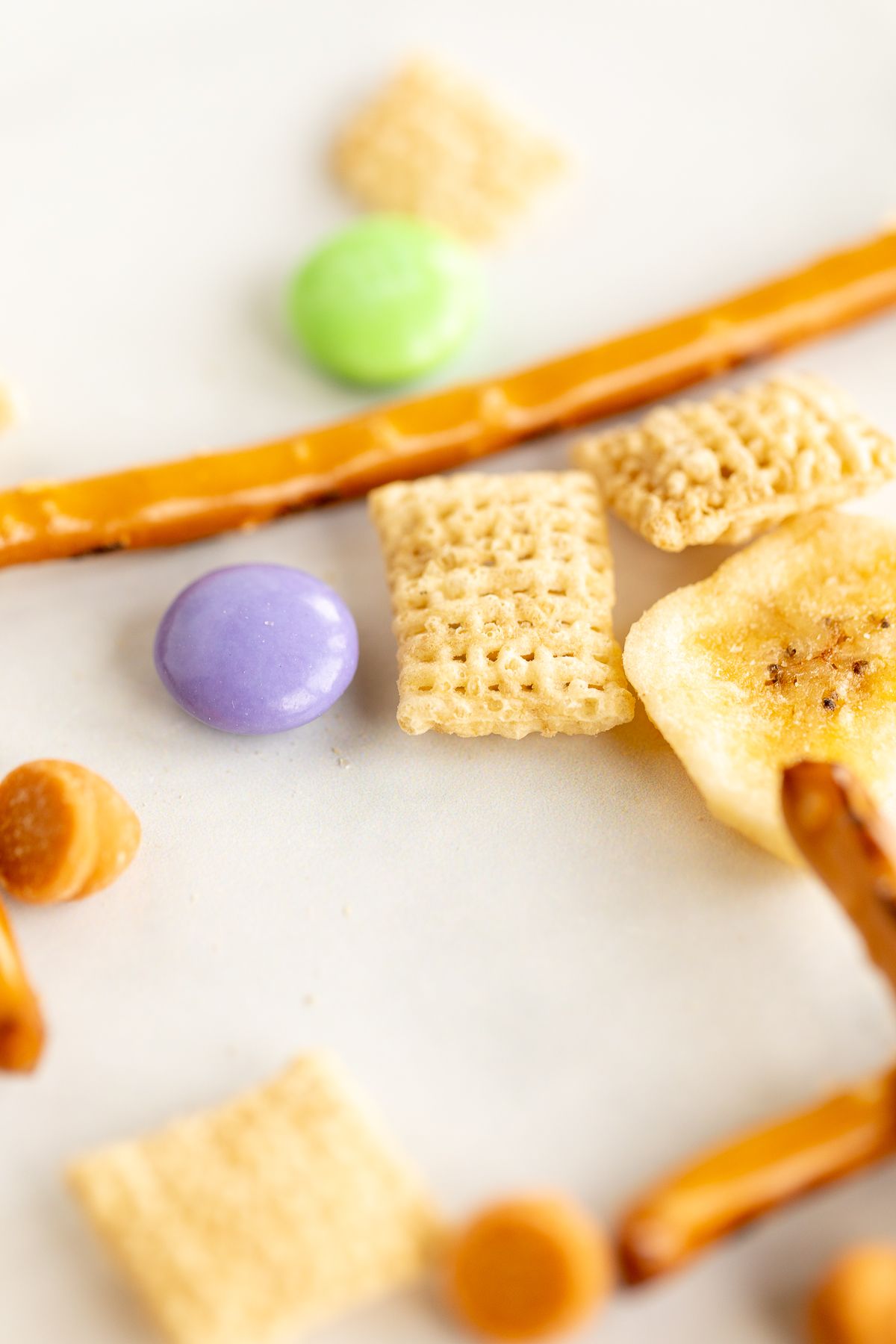 Dried banana chips, pretzels, candy and cereal on a white surface