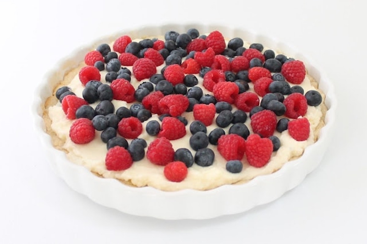 A white ceramic dish filled with a lemon berry tart, topped with fresh raspberries and blueberries on a cream base.