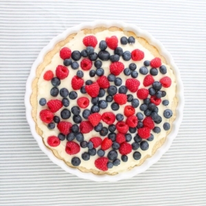 A round lemon berry tart, topped with fresh raspberries and blueberries, sits elegantly on a striped white and grey background.