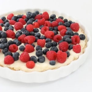 A lemon berry tart in a white dish topped with cream, blueberries, and raspberries on a white surface.