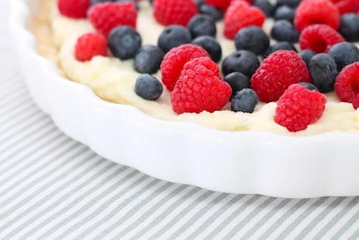 Close-up of a lemon berry tart in a white dish, topped with fresh raspberries and blueberries, placed on a light-colored, striped fabric surface.