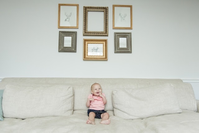 A baby sitting on a couch under a diy gallery wall.