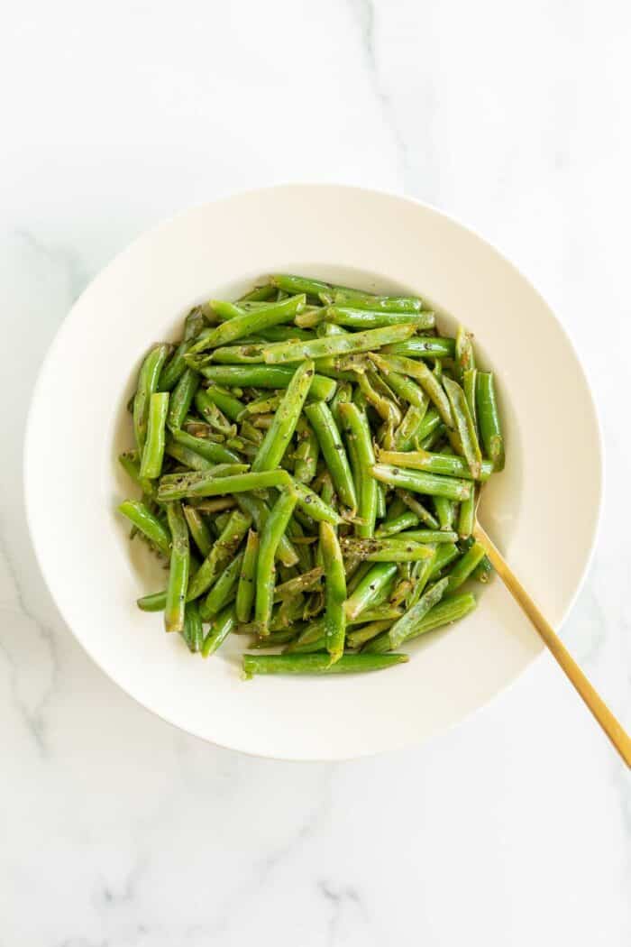 A white bowl full of seasoned green beans, gold fork for serving.