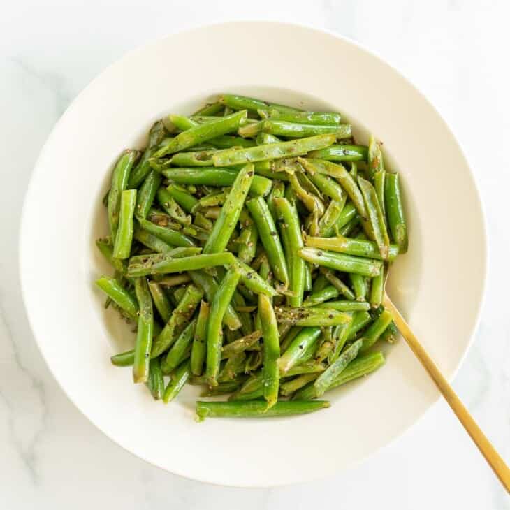 A white bowl full of seasoned green beans, gold fork for serving.