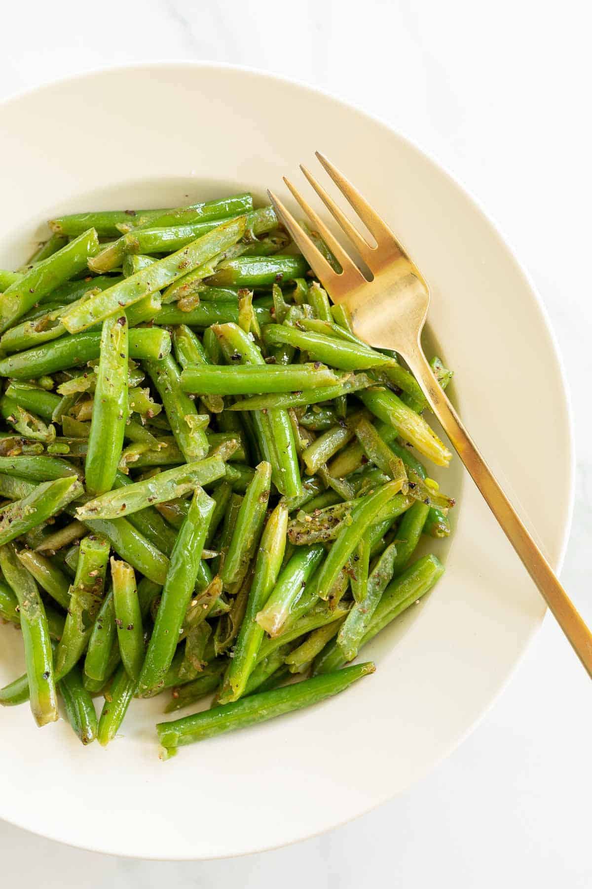 A white bowl full of seasoned green beans, gold fork for serving.