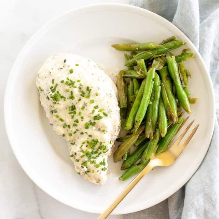 A seasoned chicken breast and seasoned green beans on a white plate with gold fork.