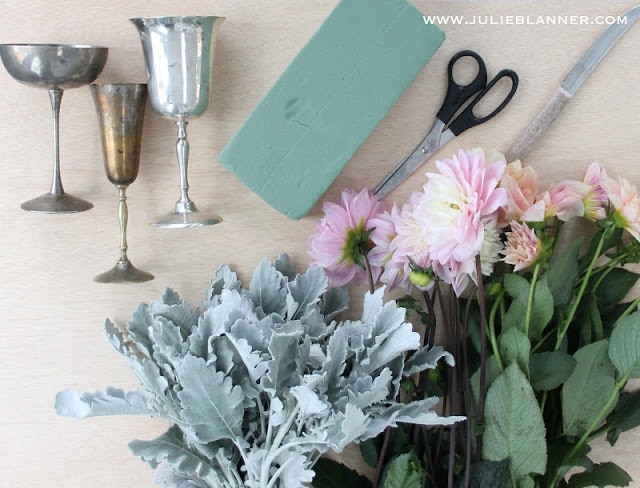 Vases, floral foam, leaves, flowers, and scissors on a wood table.