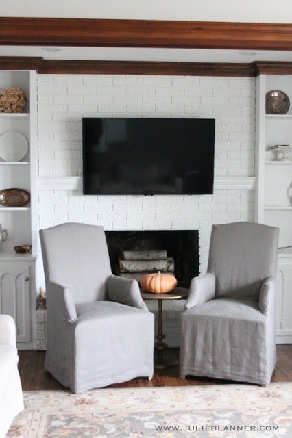 A living room with a white painted brick fireplace, with the tv handing over a mantel to hide tv cords.