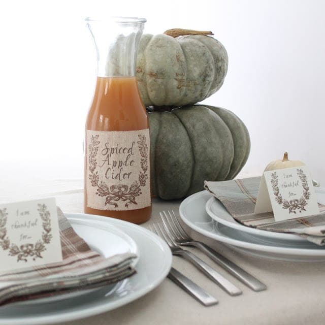Spiced apple cider in a clear glass bottle with a fabric printed label, on a table set for a fall gathering.