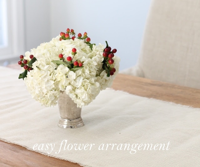 A white centerpiece with red berry accents on a white table runner, which is on a wooden table.