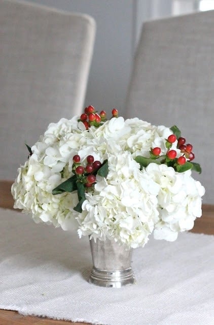 A far shot of white flowers with berry accents in a small vase on a wooden table.